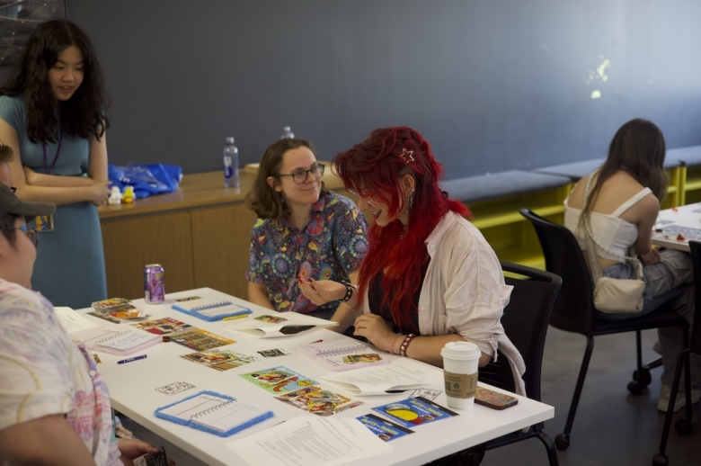 Group of Tisch Summer High school Game Design students sit at a table looking at colorful photos and notebooks during a game design class.